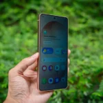 Hand holding a smartphone with green foliage background.