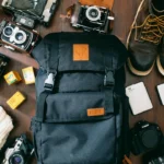 high-angle photo of black bag beside cameras, portable HDD and black leather lace-up boots